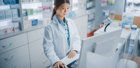 A pharmacist standing in front of a computer looks at the screen as she moves the computer’s mouse.