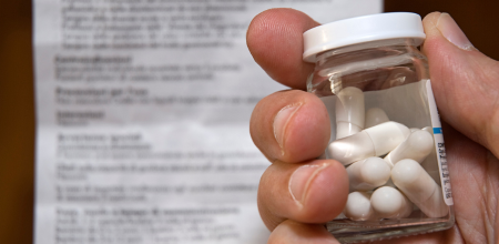 A hand holding a medicine bottle filled with tablets. A product information leaflet is blurred in the background.